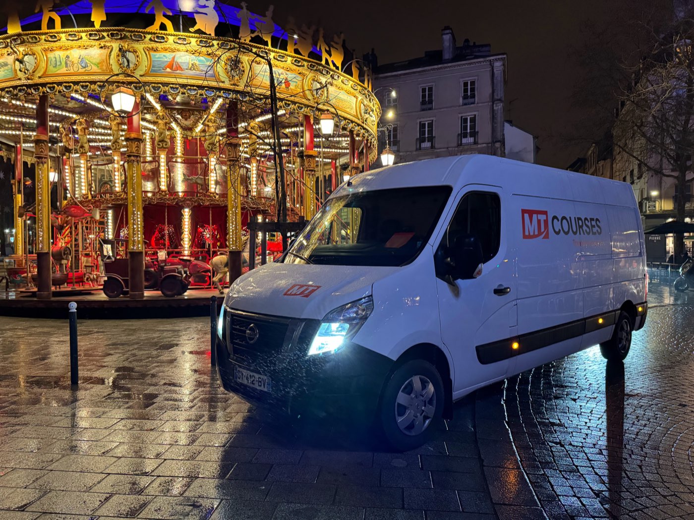 Fourgon MT Courses garé de nuit sur une place, devant un carrousel illuminé